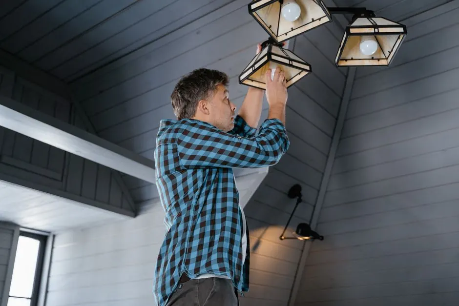 A man in a plaid shirt changes a light bulb on a ceiling lamp in a stylish, modern interior.